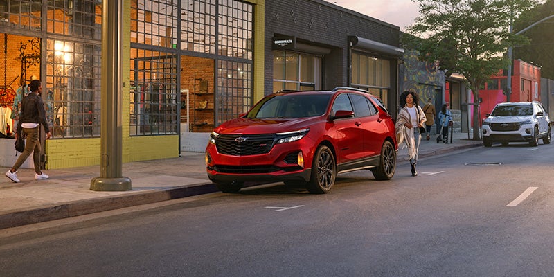 A red SUV is parked on a city street near a row of modern storefronts with large glass windows. 