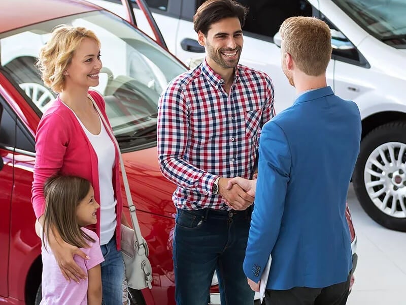 A family stands near a red car while one person shakes hands with another, likely at a car dealership.